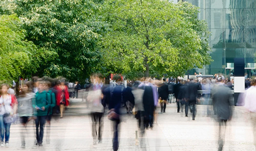 Blurred view of a busy outdoor plaza with many people walking past trees and modern buildings, capturing movement and a fast-paced urban environment.