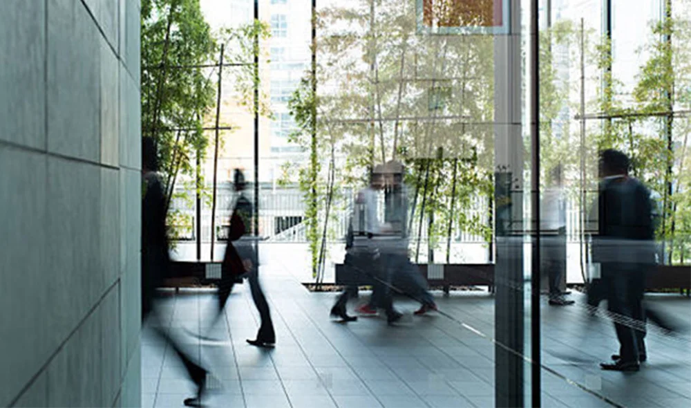 Interior of a modern office lobby with floor-to-ceiling glass walls and tall indoor plants, where several people walk past in motion blur across a polished floor, creating a sense of movement and a busy professional environment.