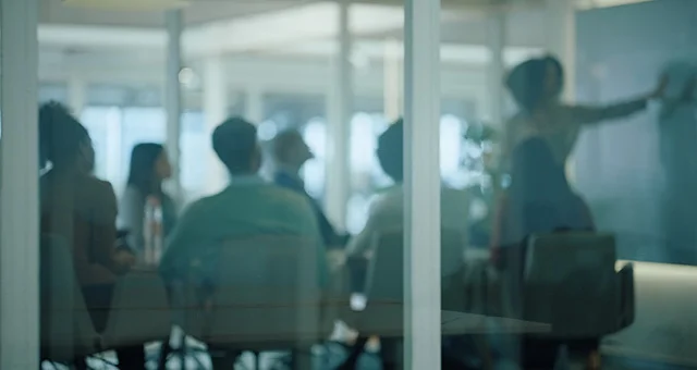 Blurry view through a glass wall of a conference room where several people are seated around a table while one person stands and gestures toward a presentation, suggesting a team meeting or discussion in a modern office setting.