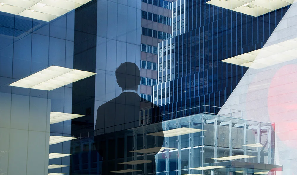 Reflection of a modern office interior with ceiling lights overlaid on glass high‑rise buildings, featuring the silhouette of a person standing and looking outward, creating a layered urban and corporate scene.