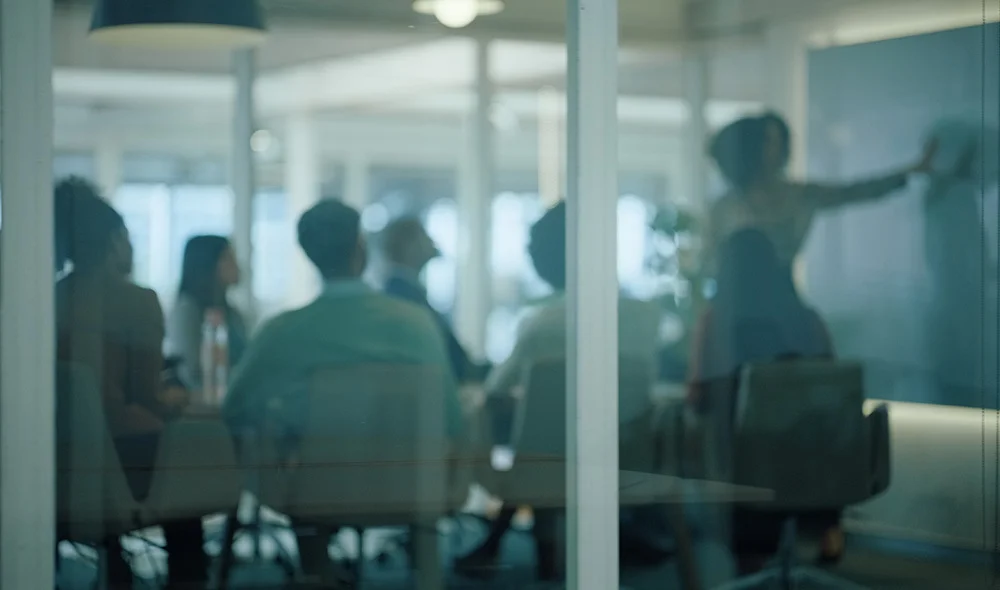 Blurry view through a glass wall of a conference room where several people are seated around a table while one person stands and gestures toward a presentation, suggesting a team meeting or discussion in a modern office setting.
