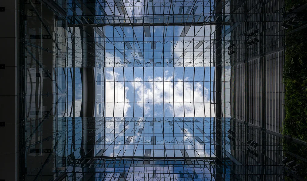 Upward view from within a modern glass atrium, showing tall reflective walls and a grid-like skylight framing blue sky and clouds, with clean architectural lines and symmetrical design.