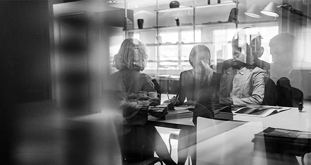 Black and white image of a group of people seated around a table in a modern office, seen through a glass partition with reflections, reviewing documents and collaborating.