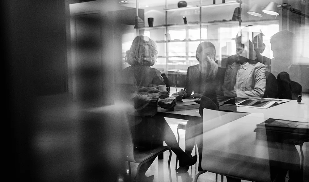 Black and white image of a group of people seated around a table in a modern office, seen through a glass partition with reflections, reviewing documents and collaborating.