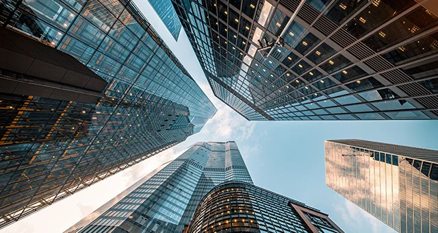 Upward view of several tall glass and steel skyscrapers from street level, with reflective surfaces and a partly cloudy sky visible between the buildings.