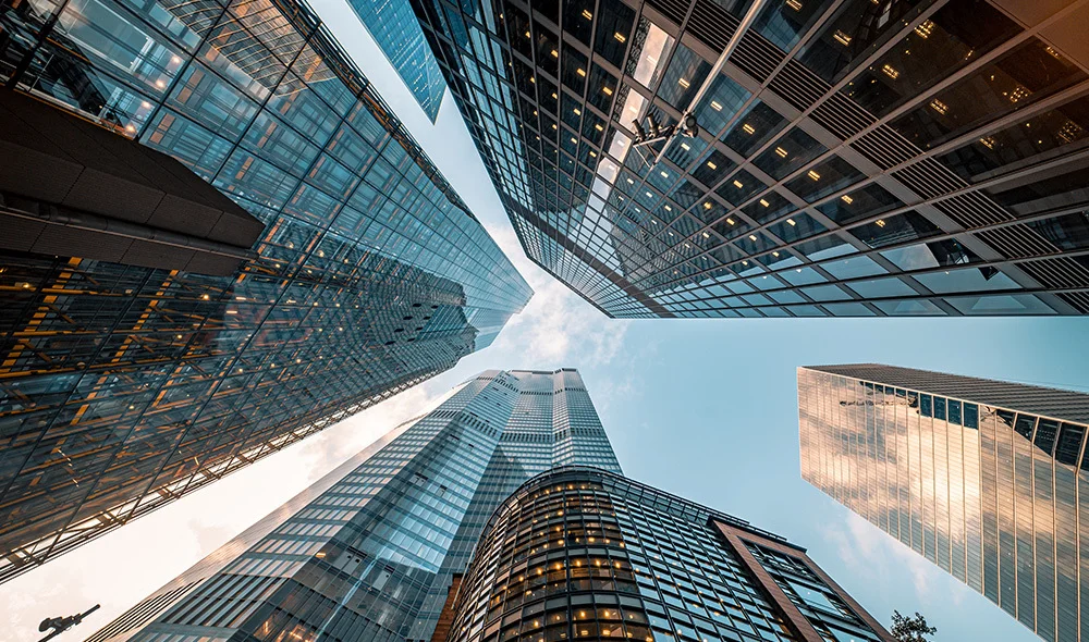 Upward view of several tall glass and steel skyscrapers from street level, with reflective surfaces and a partly cloudy sky visible between the buildings.
