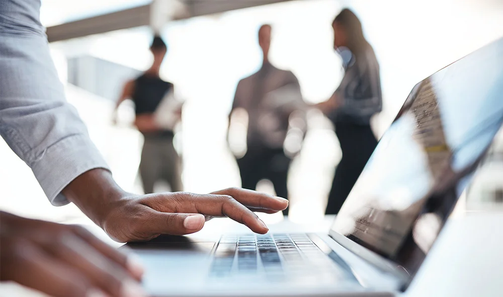 Close-up of a person’s hand using a laptop keyboard in a bright office setting, with several people standing and conversing in the blurred background, suggesting a collaborative work environment.