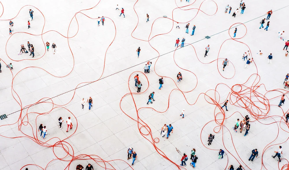 Overhead view of a large open plaza with many people spaced apart, intersected by looping red lines on the ground that weave around and encircle individuals and small groups.