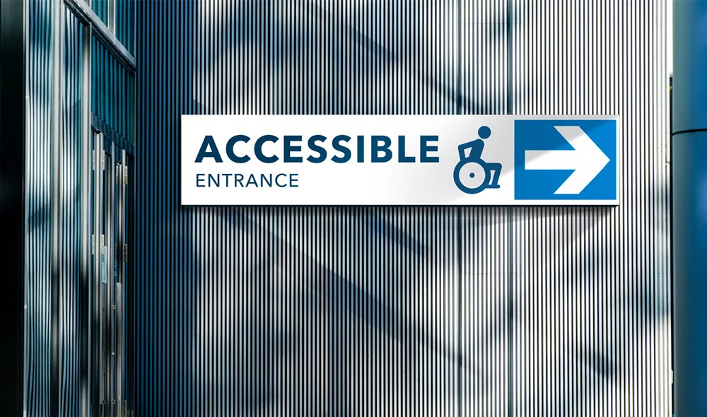 Sign reading “Accessible Entrance” with a wheelchair symbol and right-pointing arrow mounted on a modern building wall with vertical metal panels.
