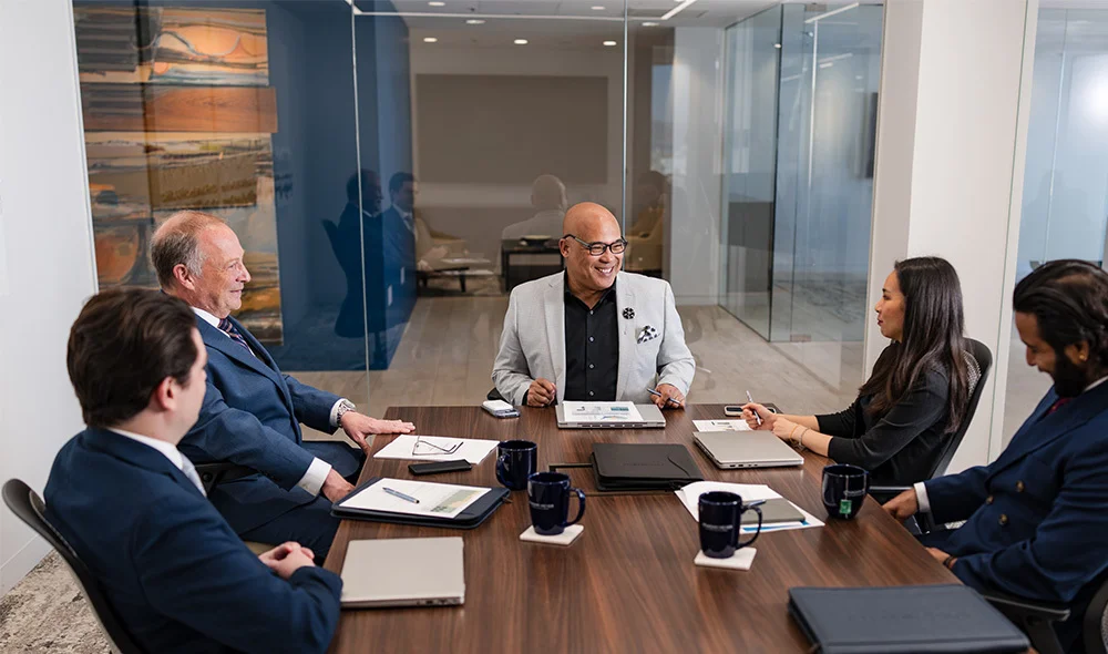 Business professionals seated around a conference table in a modern glass-walled office, reviewing documents and laptops during a meeting with coffee mugs and folders on the table.