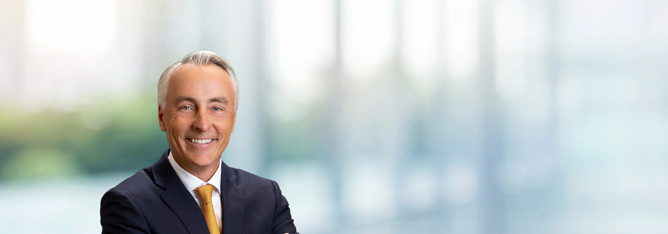 Professional headshot of Kevin O'Hagan in a dark suit and white shirt with an orange tie, facing forward with a neutral posture against a softly blurred office background with large windows and cool-toned light.