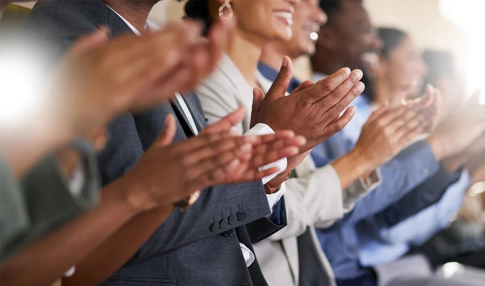 A row of attendees in business attire seated side by side, shown in a close-up as multiple hands clap in unison, suggesting applause at a formal event or presentation.