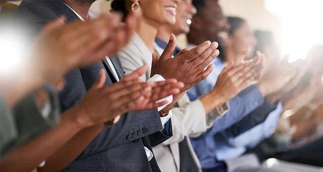 A row of attendees in business attire seated side by side, shown in a close-up as multiple hands clap in unison, suggesting applause at a formal event or presentation.