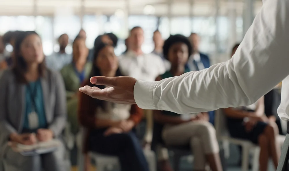A close-up view of a presenter’s outstretched hand in the foreground while a seated audience listens in a bright, modern room, suggesting a talk, workshop, or seminar.