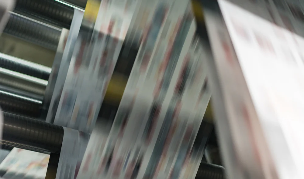 Close-up of a printing press in motion, with sheets of printed material moving rapidly through metal rollers, creating a blurred effect that conveys speed and industrial production.