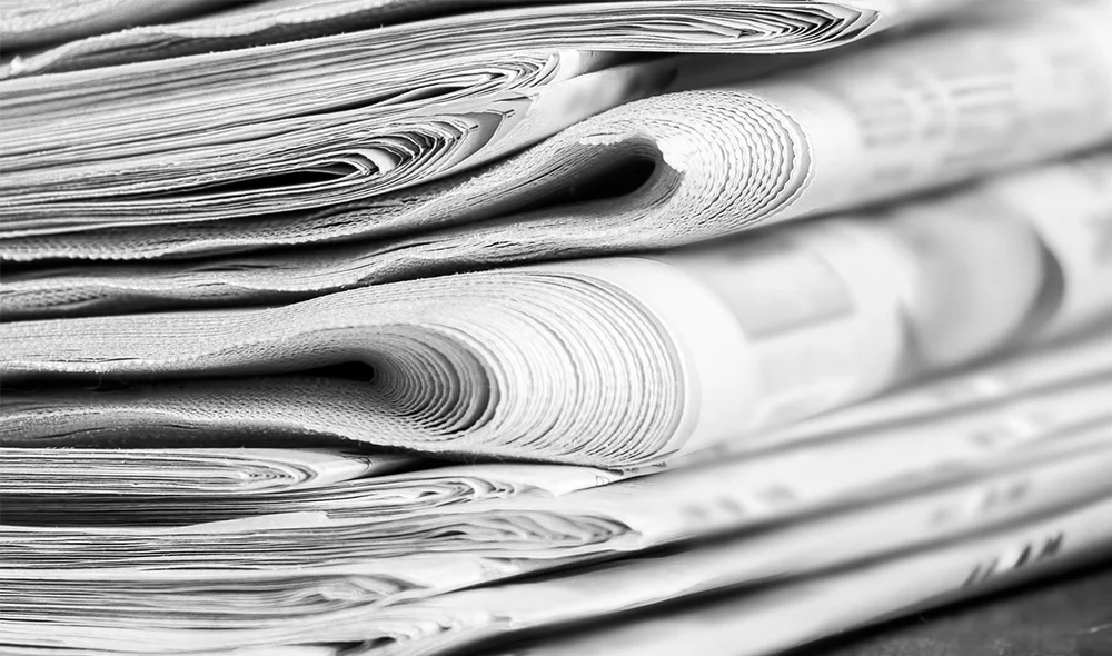 Close-up of a stack of folded newspapers in black and white, with tightly rolled edges and layered pages creating a textured, detailed pattern on a tabletop.