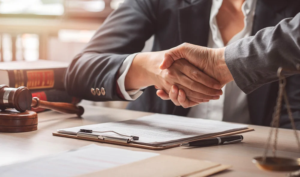 Two professionals in suits shake hands across a desk with a signed document, pen, and legal items such as a gavel and books, suggesting a business or legal agreement.