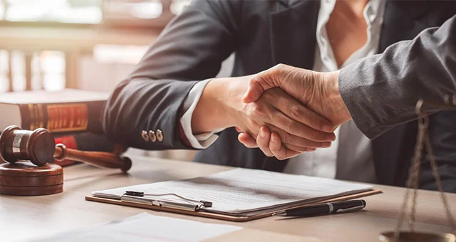 Two professionals in suits shake hands across a desk with a signed document, pen, and legal items such as a gavel and books, suggesting a business or legal agreement.
