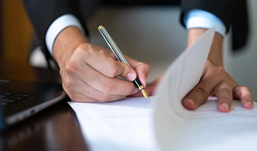 Close-up of a person in a suit signing a document with a pen while holding the pages steady on a desk, with part of a laptop visible nearby.