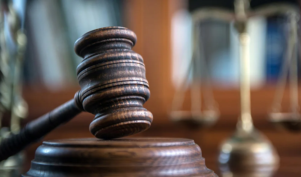 Close-up of a wooden judge’s gavel resting on a sounding block, with a blurred set of scales of justice and books in the background, representing a legal setting.