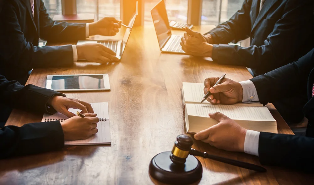 A group of professionals in suits sit around a wooden table using laptops, writing notes in notebooks, and reviewing a document, with a judge’s gavel in the foreground suggesting a legal or business meeting.