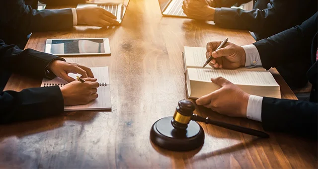 A group of professionals in suits sit around a wooden table using laptops, writing notes in notebooks, and reviewing a document, with a judge’s gavel in the foreground suggesting a legal or business meeting.