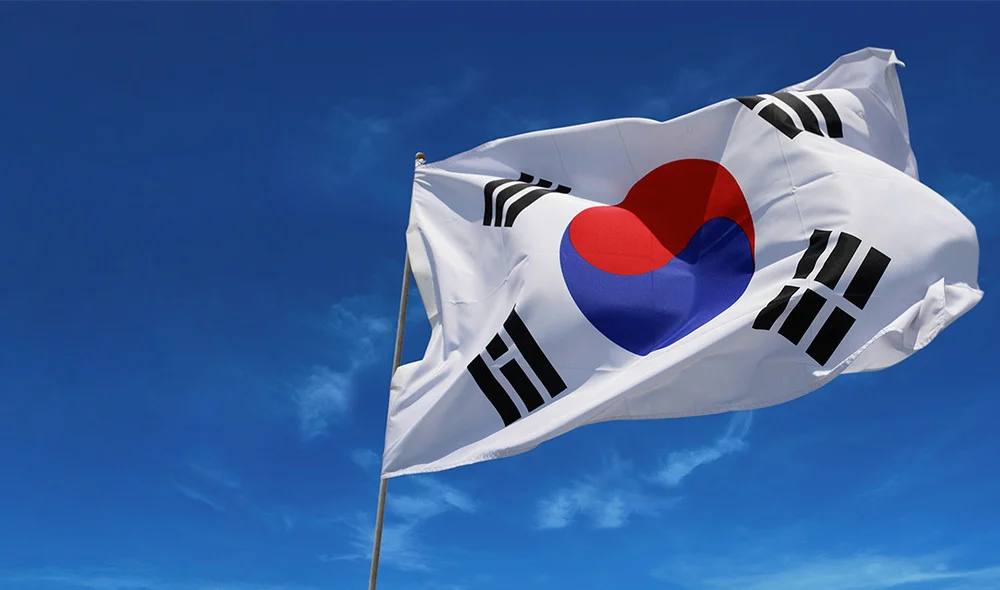 South Korean flag waving on a flagpole against a bright blue sky with light clouds. The white flag features a red-and-blue taegeuk symbol in the center and four black trigrams in each corner.