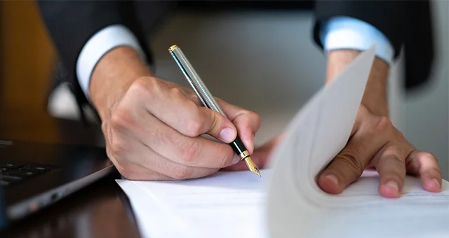 Close-up of a person in a suit signing a document with a pen while holding the pages steady on a desk, with part of a laptop visible nearby.