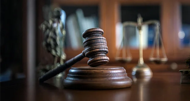 Close-up of a wooden judge’s gavel resting on a sounding block, with a blurred set of scales of justice and books in the background, representing a legal setting.